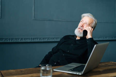 Portrait of confident gray-haired senior adult business man talking on mobile phone sitting at wooden table with laptop computer. Relaxed mature older male having conversation on smartphone.の写真素材