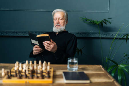 Portrait of senior gray-haired older male reading paper book at home sitting at wooden table with...