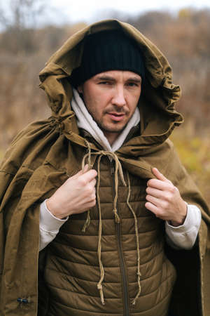Vertical portrait of serious traveler man wearing green raincoat tent standing in thicket of bushes in cold overcast day and looking at camera. Concept of survival, travel, hiking, camping in nature.の写真素材