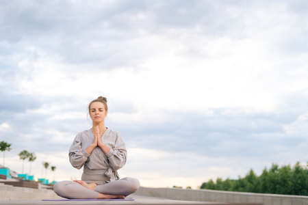 Portrait of calm Caucasian young woman practicing yoga performing namaste pose with closed eyes outside in city park. Pretty serene female sitting lotus position on yoga mat outdoors alone.の写真素材