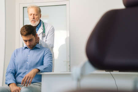 Front view of mature adult male manual therapist turning and bending head of male patient to check...