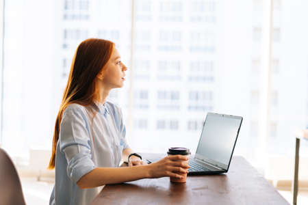 Side view of thoughtful young business woman sitting at desk with laptop in modern office room,...