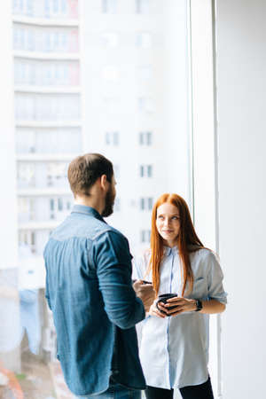 Vertical shot of young startup man and woman coworkers with coffee cups standing by window in loft...