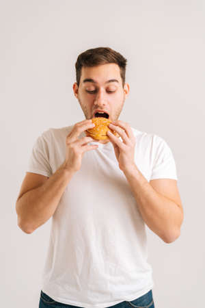 Vertical shot of handsome young man with enjoying eating delicious burger on white isolated background. Studio portrait of happy handsome male holding tasty unhealthy hamburger.の写真素材