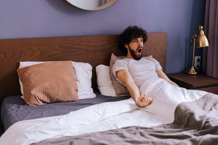 Handsome bearded young man with open mouth waking up stretching arms lying in bed on early morning on holiday. Relaxed male enjoying wake-up in bedroom at weekend, wants to stay in bed for long time.の写真素材