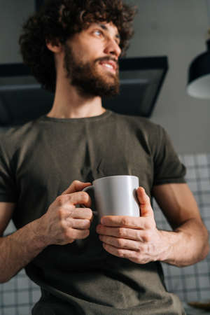 Close-up low-angle view of pensive young man holding in hand cup with morning coffee, thoughtful looking away, standing in kitchen with modern light interior, enjoying fresh warm drink at home office.の写真素材
