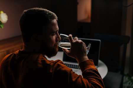 Back view of tired young man drinking wine during working on laptop at nighttime sitting at table in dark room. Bearded male sitting at home and drinking wine while browsing internet on computer.の写真素材