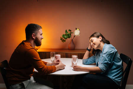 Side view of happy young couple enjoying talking, having fun together celebrating Valentines day dining during romantic dinner date, sitting together at table with candles in cozy dark living room.の写真素材