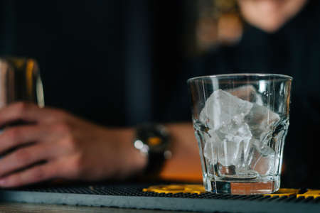 Close-up cropped shot of unrecognizable bartender male standing behind bar counter, glass filled with ice cubes on foreground. Barman making cocktail with ice in modern bar with dark interior.の写真素材