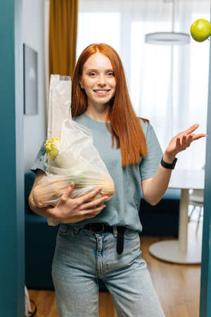 Vertical portrait of pretty young woman housewife holding groceries bag and smiling looking at...