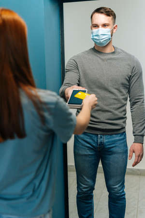 Vertical shot of handsome courier male in protective face mask giving POS wireless terminal to making contactless payment via credit card, to unrecognizable female customer on doorstep at aparment.の写真素材