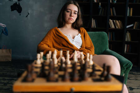 Portrait of thoughtful young woman in elegant eyeglasses thinking about chess move sitting in...
