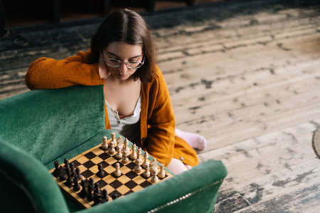 High-angle view of attractive young woman wearing elegant eyeglasses thinking about chess move while sitting on wooden floor, by armchair in dark room playing logical board game alone.の写真素材