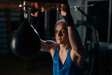 Portrait of brutal bald boxer male in boxing defence tape resting near punching bag in sport club with dark interior, looking at camera. Tired fighter relaxing after training.の写真素材