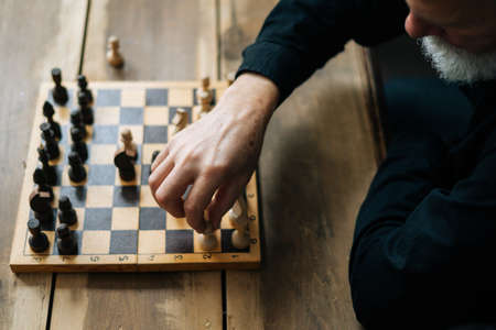 Close-up top view of unrecognizable senior bearded chess player performing move with pawn piece on wooden chessboard. Closeup of businessman making move with white pawn on chess board.の写真素材