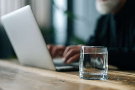 Close-up cropped shot of unrecognizable mature male using laptop sitting at table in dark room, selective focus, blurred background. Closeup of aged business man working on computer indoors.の写真素材