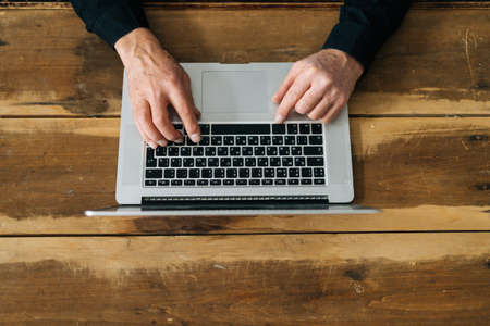 Close-up top view of unrecognizable senior male using typing on laptop keyboard sitting at wooden...