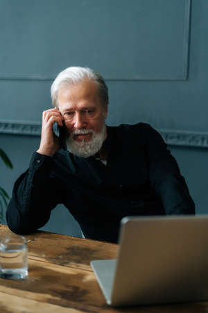 Vertical portrait of serious gray-haired mature aged businessman talking on mobile phone sitting at table with laptop computer at home office. Senior older male having conversation on smartphone .の写真素材