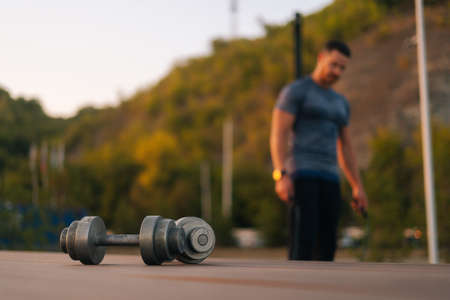 Blurred unrecognizable sportsman with strong muscular body looking at camera during summer evening workout outdoors. Focus on foreground to dumbbells, selective focus.の写真素材