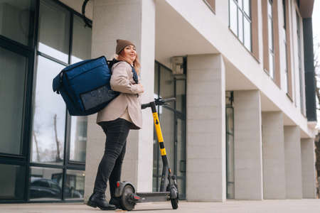 Low-angle view of positive female courier with big thermo backpack standing with electric scooter in...