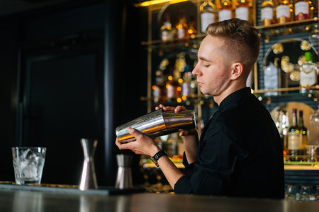 Confident barman in black clothes mixing ingredients of alcoholic cocktail by shaking shaker standing behind bar counter, on blurred background of shelves with alcoholic drinks, selective focus.の写真素材