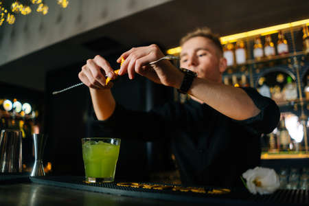 Close-up low-angle view of young bartender male squeezing out piece of orange peel with straw and decorating cocktail, standing behind bar counter in modern nightclub with dark interior.の写真素材