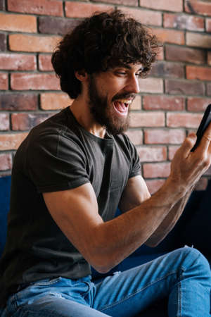 Vertical shot of happy excited young man looking at mobile phone screen, reading message with good...