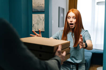 Surprised young woman receiving paper boxes with hot pizza from unrecognizable courier male on doorway at home. Rear view of delivery man delivering boxes with food to female client at apartment.の写真素材