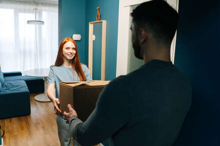 Smiling redhead young woman receiving cardboard box from unrecognizable delivery man on doorstep at apartment. Rear view of courier male delivering parcel package post to female client at home.の写真素材