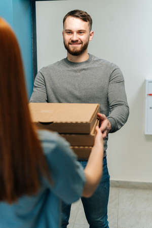 Vertical shot of bearded courier male delivering boxes with hot pizza to unrecognizable female customer on doorway at home. Rear view of young woman client receiving boxes with food from delivery man.の写真素材