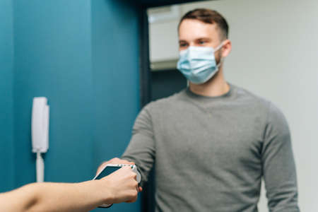 Selective focus of courier male wearing protective face mask giving POS wireless terminal to making contactless payment via credit card, to unrecognizable female customer on doorstep at home.の写真素材