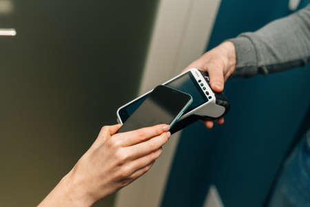 Close-up high-angle view of unrecognizable courier male giving POS wireless terminal to making contactless payment using smartphone to young female customer, on doorstep at apartment.の写真素材