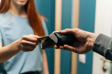 Close-up cropped shot of unrecognizable courier male giving POS wireless terminal to making contactless payment via credit card to young female customer, on doorway at home.の写真素材