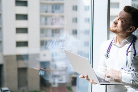 Blinking from sun male doctor in white uniform with stethoscope using laptop holding with one hand, standing near window in sunny day. Concept of modern medicine and healthcare.の写真素材