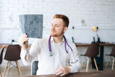 Portrait of focused male doctor wearing white uniform examining brain computerized tomography scan sitting at desk in hospital office, thinking about diagnosis. Concept of medicine and healthcare.の写真素材