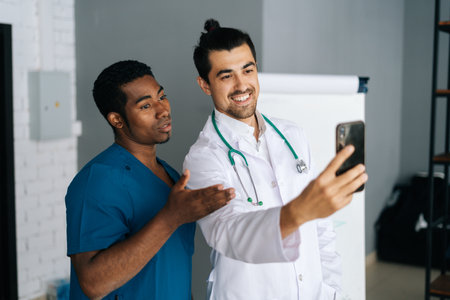 Cheerful African and Caucasian men in medical uniform taking selfie on smartphone standing in hospital conference office. Two happy medical workers making photo after international meeting.の写真素材