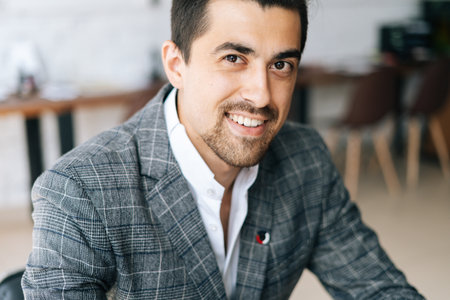 Close-up face of cheerful elegant business man wearing fashion suit sitting at desk in modern office room, looking at camera. Front view of serious bearded businessman posing at workplace.の写真素材