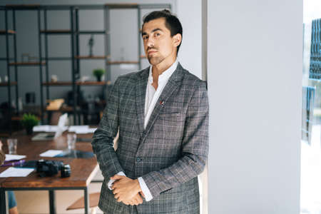 Portrait of confident young business man wearing stylish suit standing near window in modern office room, looking at camera. Front view of handsome bearded businessman posing at workplace.の写真素材