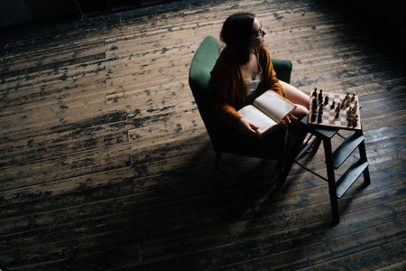 High-angle view of thoughtful young woman in elegant eyeglasses holding book sitting in armchair in dark vintage room, thinking looking away. Serious businesswoman planning development strategy.の写真素材