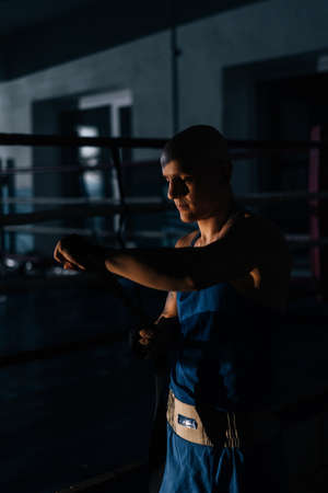 Vertical shot of confident boxer wrapping defense boxing tape around arms before fight in sport club with dark interior on background of ring. Serious fighter getting ready for fight in fitness centerの写真素材
