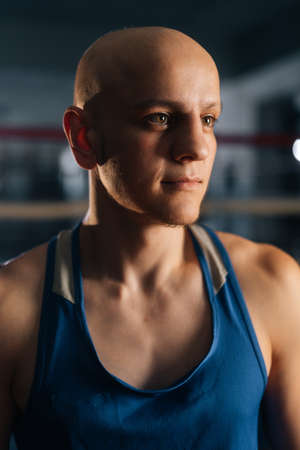 Close-up vertical shot of professional bald brutal boxer wearing sportswear standing posing in sport club with dark interior, looking away. Closeup view of young fighter training in gym.の写真素材