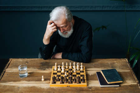 High-angle view of thoughtful bearded mature male thinking about chess move sitting at home and...