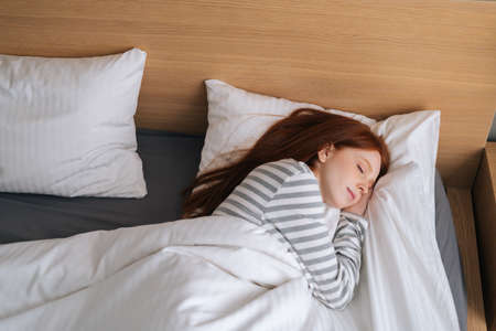 Top view of pretty redhead young woman sleeping well in bed hugging soft white blanket at home. Charming lady resting enjoying fresh soft bedding linen and mattress in bedroom.の写真素材
