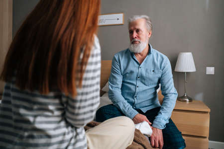 Back view of loving young granddaughter talking with gray-haired grandfather sitting in bed at cozy bedroom. Happy young woman and mature male having fun, enjoying leisure time, sharing news.の写真素材