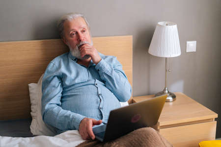 High-angle view of thinking gray-haired senior male with beard using laptop computer sitting on bed at home. Pensive bearded mature older man in pajamas having online video call via notebook.の写真素材