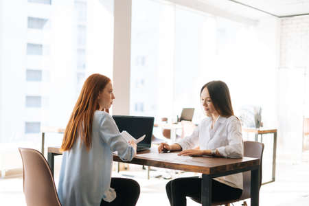 Back view of two young business women discussing financial data in report using laptop and paper...