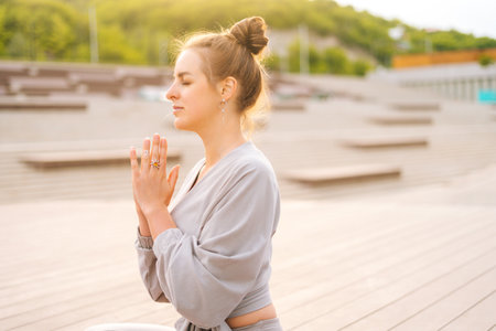 Side view of serene Caucasian young woman practicing yoga performing namaste pose with closed eyes outside in city park. Pretty calm female sitting lotus position on yoga mat outdoors alone.の写真素材