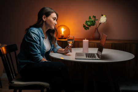 Attractive young woman enjoying distant dating using laptop, holding glass with champagne, talking celebration toasting via video call looking on screen. Couple in love chatting on remote date.の写真素材