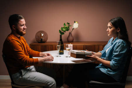 Bearded loving man giving box with gift to charming young woman sitting at table with candles on...