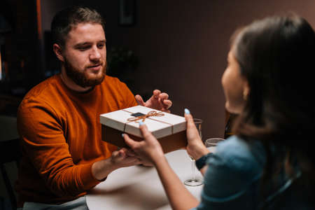 Happy young man receiving present from loving wife enjoying romantic dinner date sitting at table with candles on birthday or Valentines Day. Pretty young woman giving wrapped box with gift to husbandの写真素材